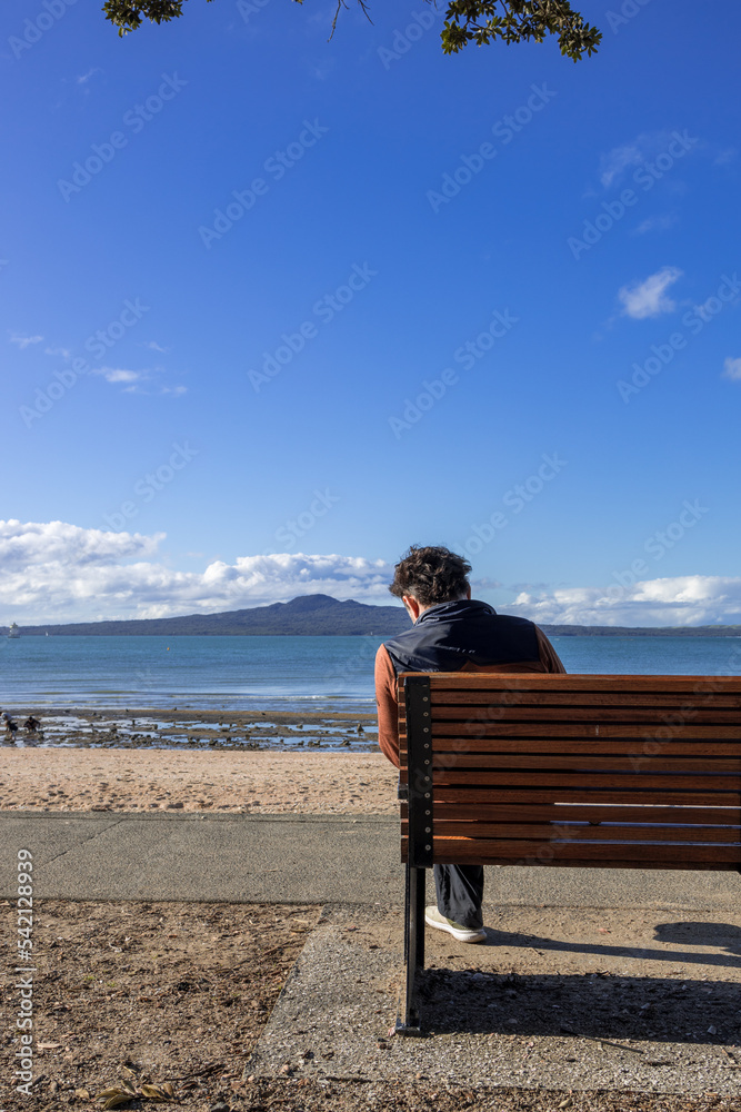 Young adult male sitting on the bench facing the sea at Mission Bay Beach, Auckland New Zealand NZ Young adult male sitting on the bench facing the sea at Mission Bay Beach, Auckland New Zealand NZ