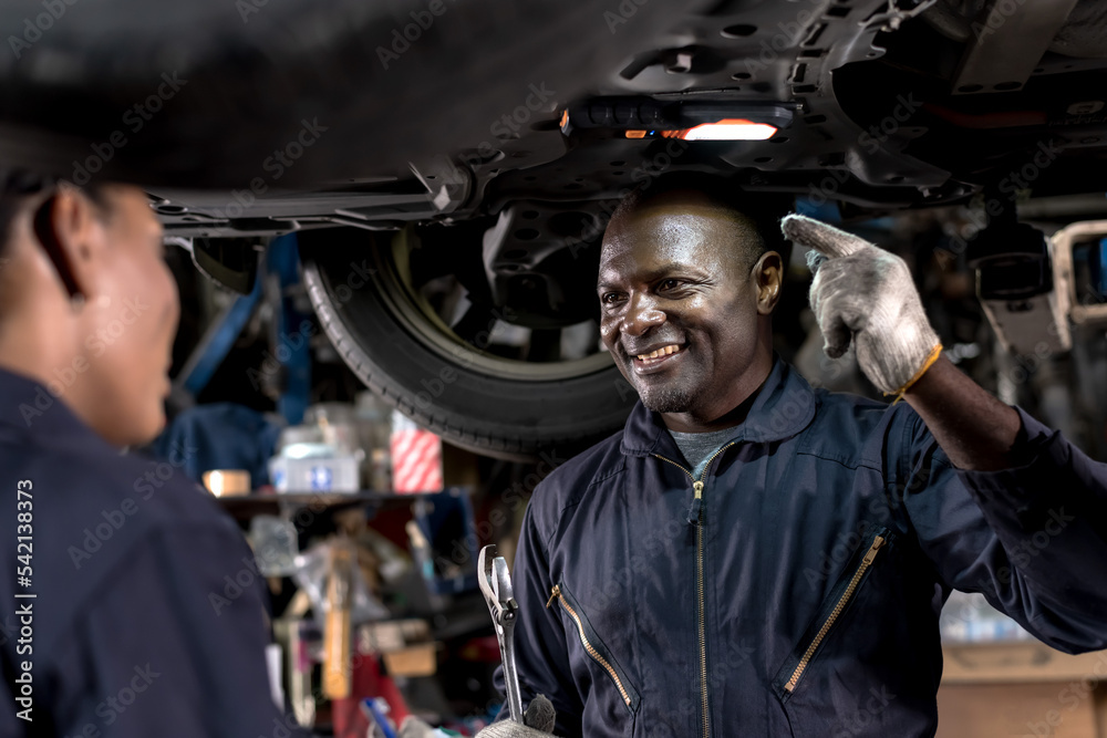 Smiling black African mechanic man in overall uniform working underneath lifted vehicle in auto repair shop,  Car Mechanic Concept