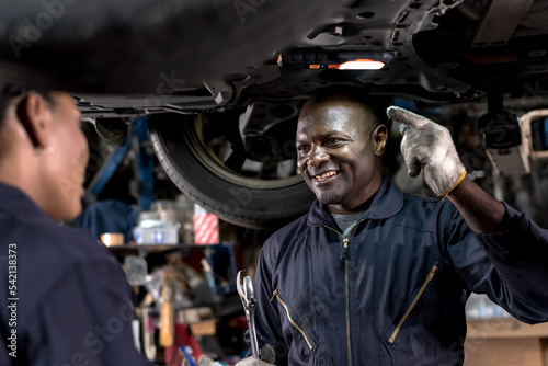 Smiling black African mechanic man in overall uniform working underneath lifted vehicle in auto repair shop, Car Mechanic Concept