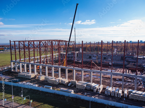 Aerial view of the sports complex under construction. Construction cranes lift the beams of the metal frame of the building onto a concrete base.