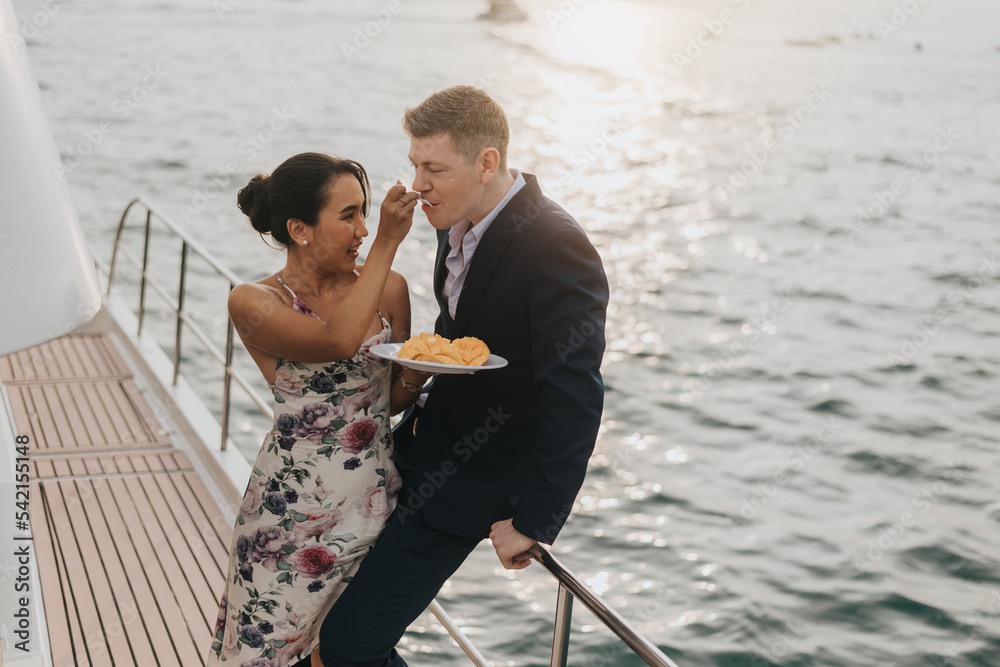 Romantic couple love eating on yacht. Happy woman standing and giving fruit to her husband in cruise ship while sunset on vacation. luxury and honeymoon lifestyle, Happy aniversary.