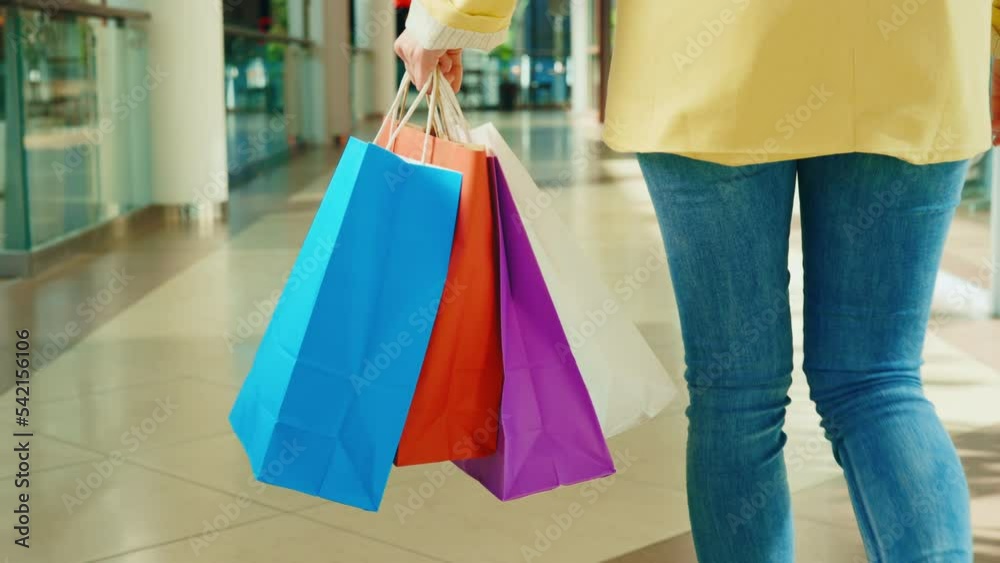 Back close view of the woman going on the mall, holding in her hands many colorful paper packages from the shop. Woman after shopping
