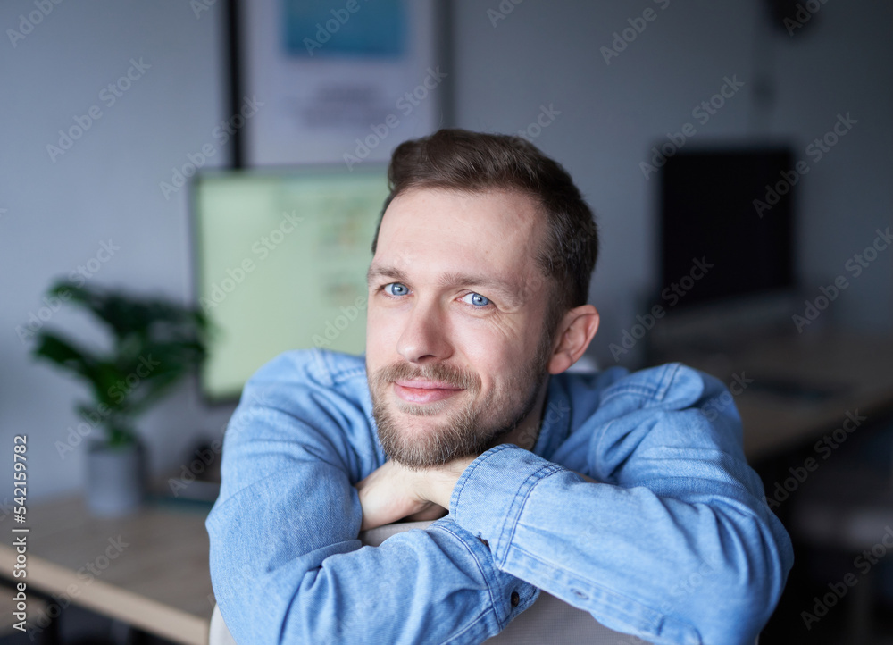 Obraz premium Attractive bearded male sitting in home office looking at camera smiling. Young handsome man in denim shirt, positive emotion with computer monitor on background. High quality image