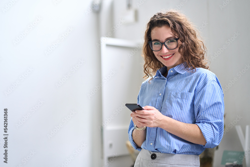 Smiling young business woman user, happy businesswoman wearing glasses holding cellular smartphone working standing in office using mobile cell phone working on cellphone looking at camera. - obrazy, fototapety, plakaty 