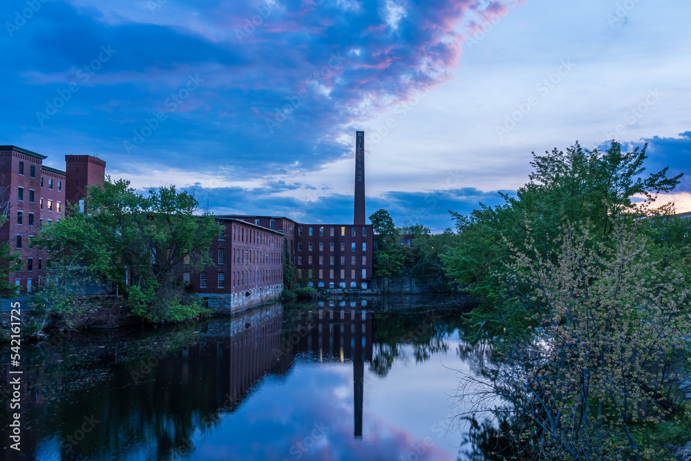 Historic cotton mill buildings with a tall brick chimney in an old industrial park with reflection in the river in the late evening. Nashua, New Hampshire, USA