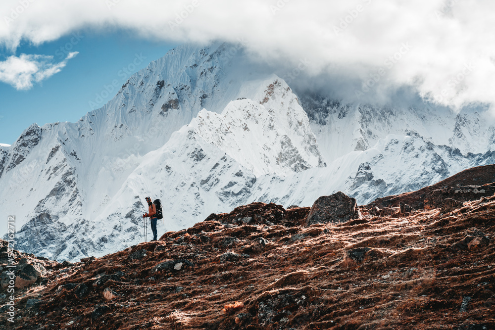 Solo tourist with travel backpack stand in front of snowy and cloudy mountains. Traveler among high altitude mountain