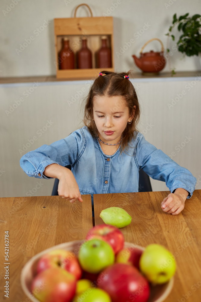 Fototapeta premium Happy little girl twist the pear, the bowl with variety fruits on table at home