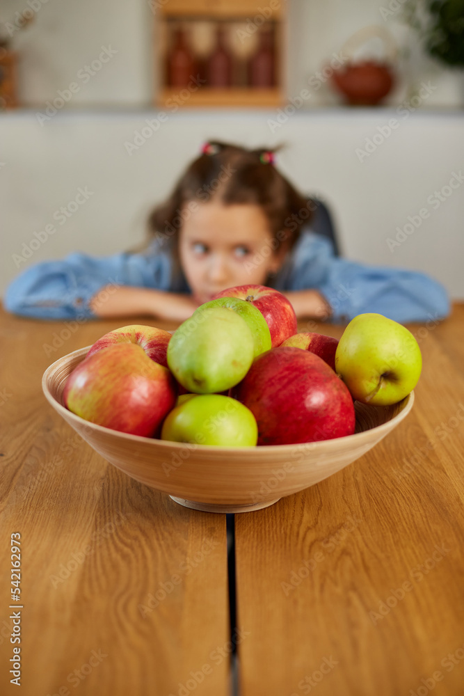 Fototapeta premium Little sad girl look on bowl with fruits in the kitchen at home