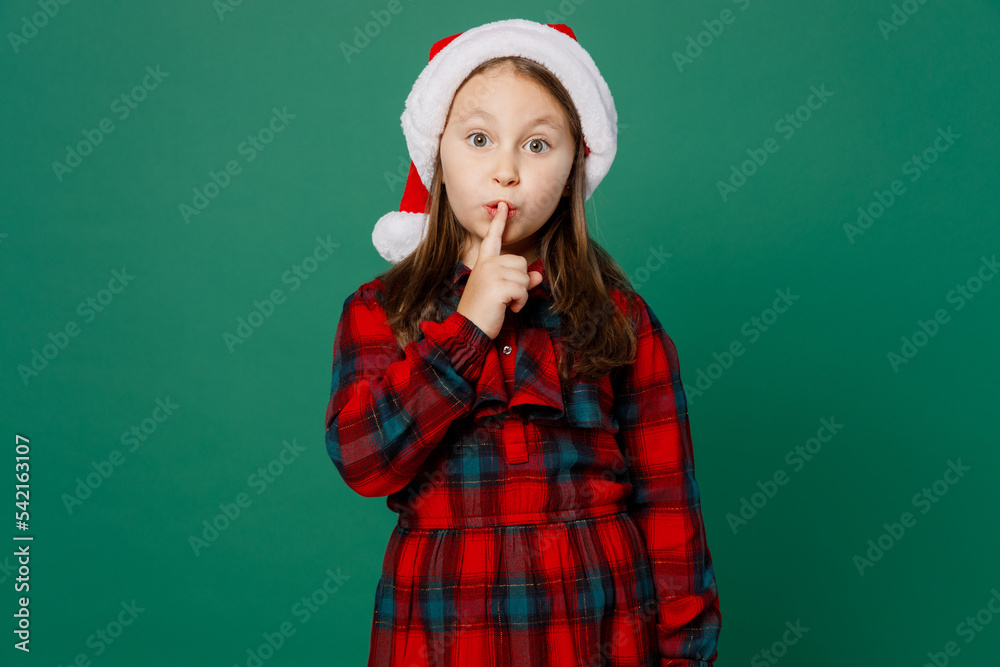 Merry secret little child kid girl 7 years old wear red dress Christmas hat posing say hush be quiet with finger on lips shhh gesture isolated on plain green background. Happy New Year 2023 concept. Merry secret little child kid girl 7 years old wear red dress Christmas hat posing say hush be quiet with finger on lips shhh gesture isolated on plain green background. Happy New Year 2023 concept.
