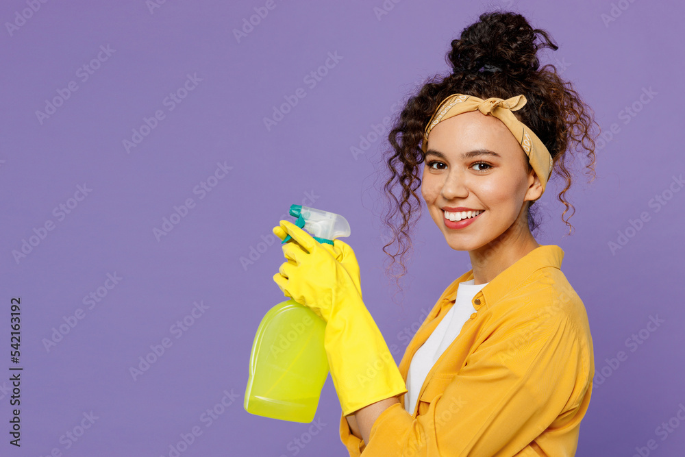 Side view happy young housekeeper woman in yellow shirt tidy up hold in hand bottle use spray detergent cleanser look camera isolated on plain pastel light purple background studio Housework concept Side view happy young housekeeper woman in yellow shirt tidy up hold in hand bottle use spray detergent cleanser look camera isolated on plain pastel light purple background studio Housework concept