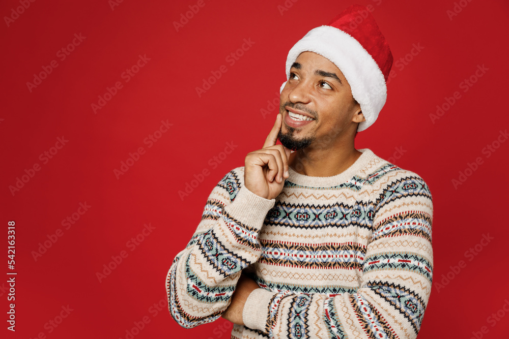 Merry young man wear warm Christmas sweater Santa hat posingl look aside on workspace area mock up prop up chin isolated on plain red color background. Happy New Year 2023 celebration holiday concept. Merry young man wear warm Christmas sweater Santa hat posingl look aside on workspace area mock up prop up chin isolated on plain red color background. Happy New Year 2023 celebration holiday concept.