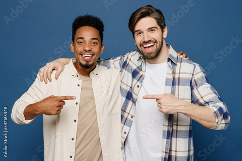 Young two friends fun cool men wear white casual shirts together looking camera put hand on shoulder point finger on each other isolated plain dark royal navy blue background People lifestyle concept photo