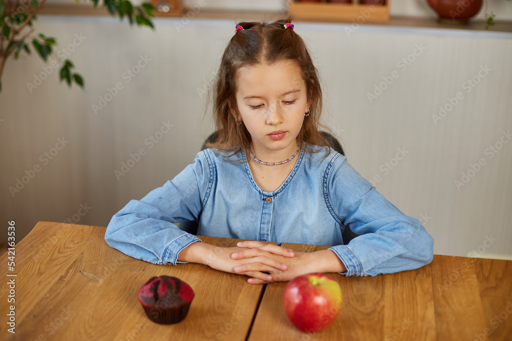 Fototapeta premium Little girl looking on sweet cake and fresh red apple and choosing in kitchen