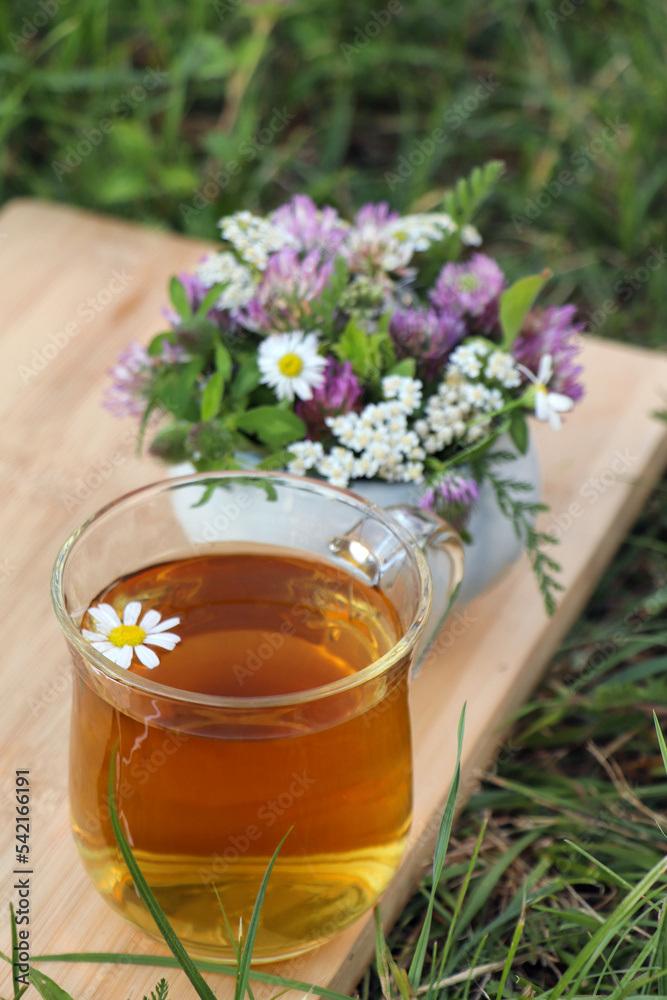 Cup of aromatic herbal tea and ceramic mortar with different wildflowers on green grass outdoors