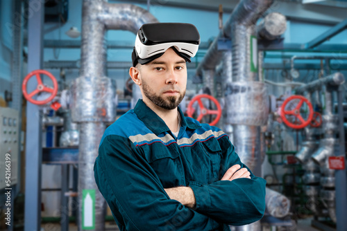 Modern industry and manufacturing. Caucasian young serious man worker in uniform, hardhat and vr glasses, arms crossed against background of industrial boilers. Engineering and energy concept © _KUBE_