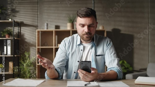 Young man using cellphone software applications sitting at table, feeling dissatisfied irritated by bad device work, poor internet connection