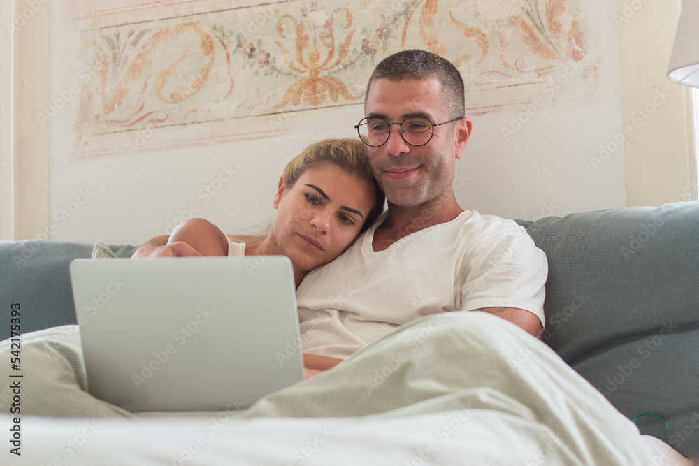 Cute man and woman hugging in bed and looking at laptop screen. Cheerful young couple spending morning together, relaxing and watching movie online. Love, sleep cycle, family concept