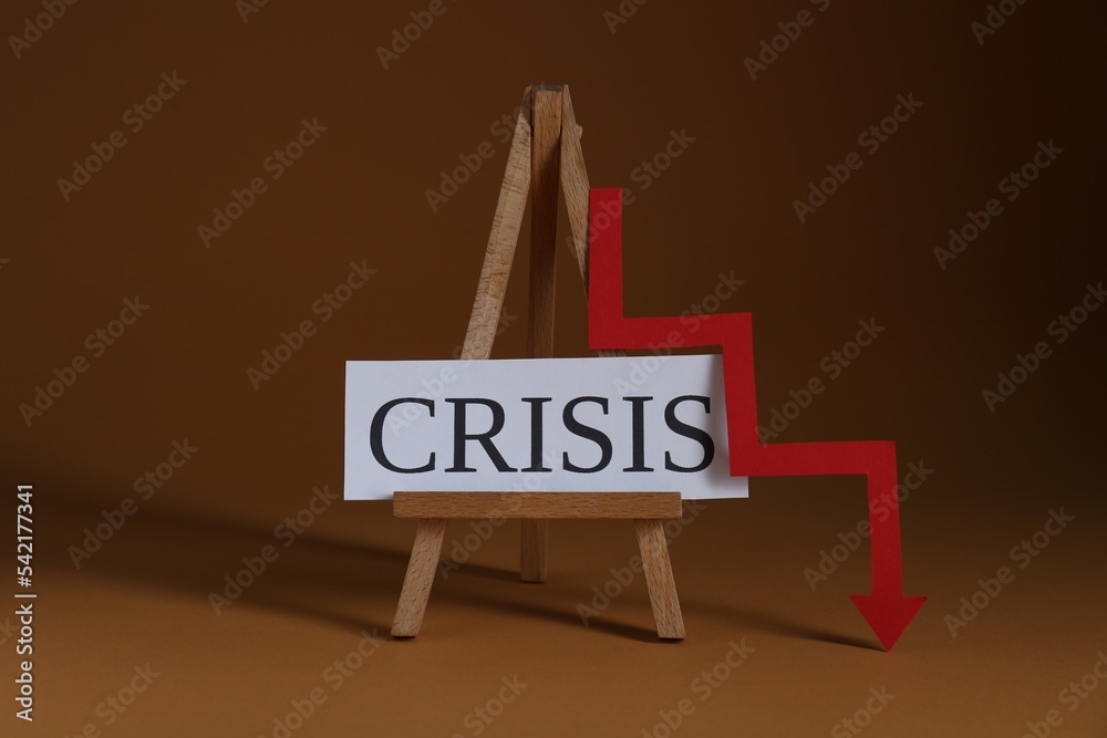 Wooden easel with word Crisis and descending red arrow on brown background Wooden easel with word Crisis and descending red arrow on brown background