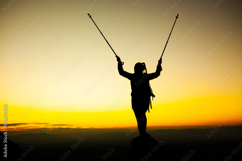 Silhouette of a hiker girl on a rock pedestal with hands up. Beautiful orange sunset.