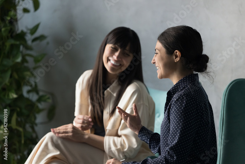 Cheerful multi ethnic women laughing seated indoor. Middle-aged and millennial workmates having friendly talk, joking at break meet in workspace, discuss project, share creative ideas feel satisfied