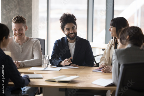 Smiling 35s Indian male boss, team leader talking to clients or corporate staff at briefing seated at table in office, businessman executive negotiating at diverse group, take part in formal meeting