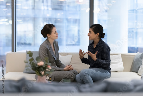 Multiethnic businesswomen, Caucasian and Indian female workmates sit on cozy sofa in modern office lobby lead informal conversation during break at workspace. Communication at workplace, discussion