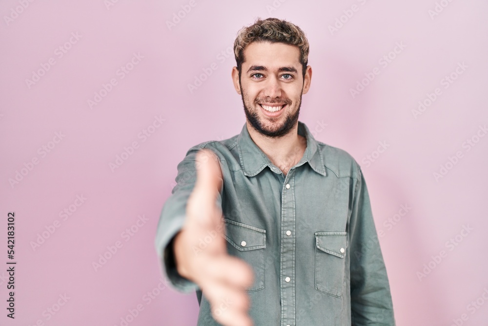 Hispanic man with beard standing over pink background smiling friendly offering handshake as greeting and welcoming. successful business. Hispanic man with beard standing over pink background smiling friendly offering handshake as greeting and welcoming. successful business.