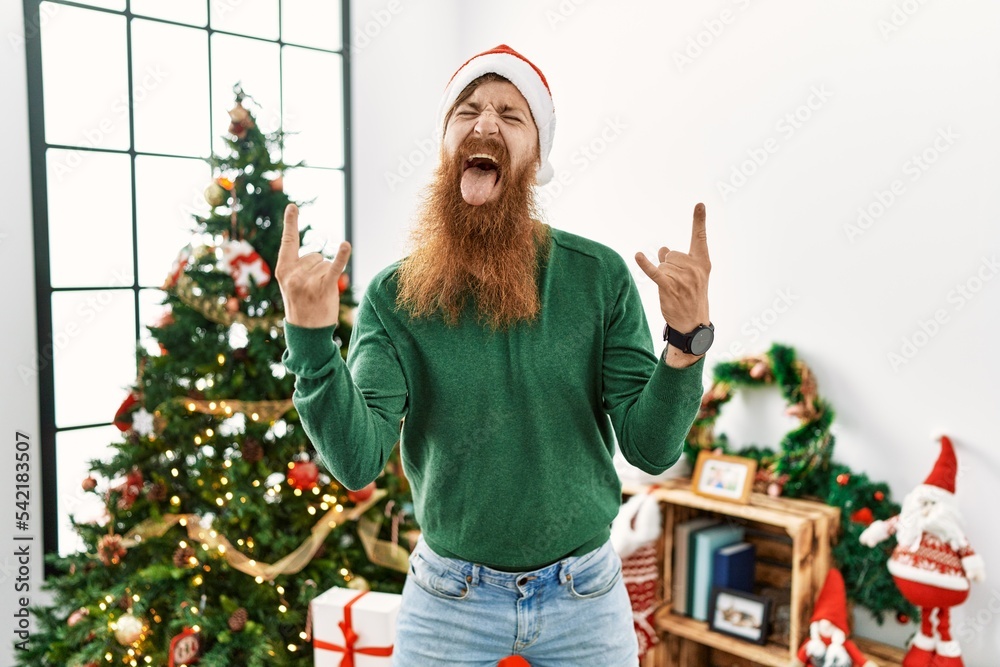 Redhead man with long beard wearing christmas hat by christmas tree shouting with crazy expression doing rock symbol with hands up. music star. heavy music concept. Redhead man with long beard wearing christmas hat by christmas tree shouting with crazy expression doing rock symbol with hands up. music star. heavy music concept.