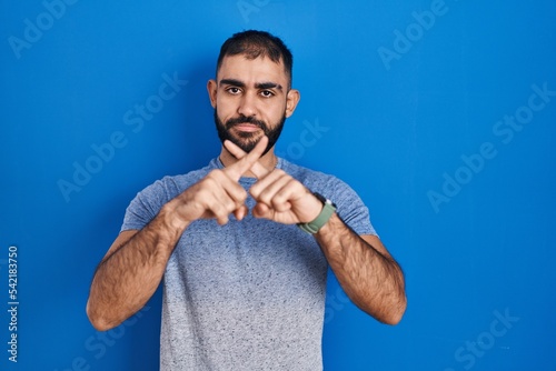 Middle east man with beard standing over blue background rejection expression crossing fingers doing negative sign