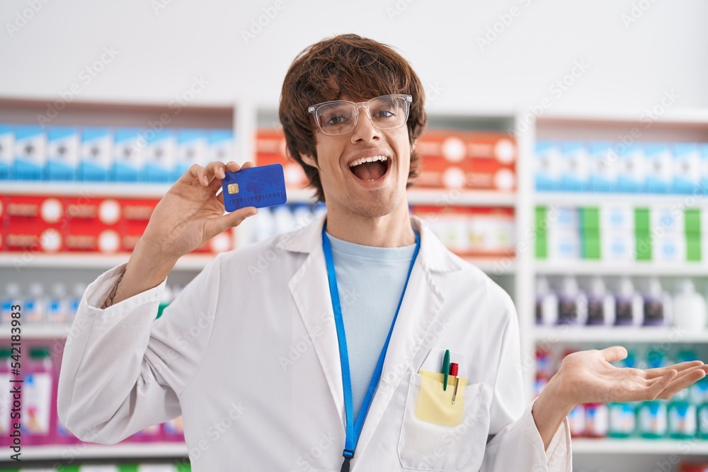 Hispanic young man working at pharmacy drugstore holding credit card celebrating achievement with happy smile and winner expression with raised hand Hispanic young man working at pharmacy drugstore holding credit card celebrating achievement with happy smile and winner expression with raised hand