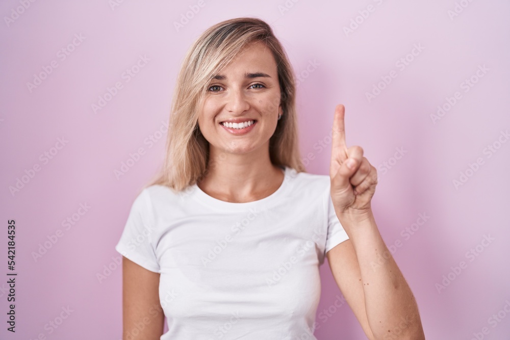 Young blonde woman standing over pink background showing and pointing up with finger number one while smiling confident and happy. Young blonde woman standing over pink background showing and pointing up with finger number one while smiling confident and happy.
