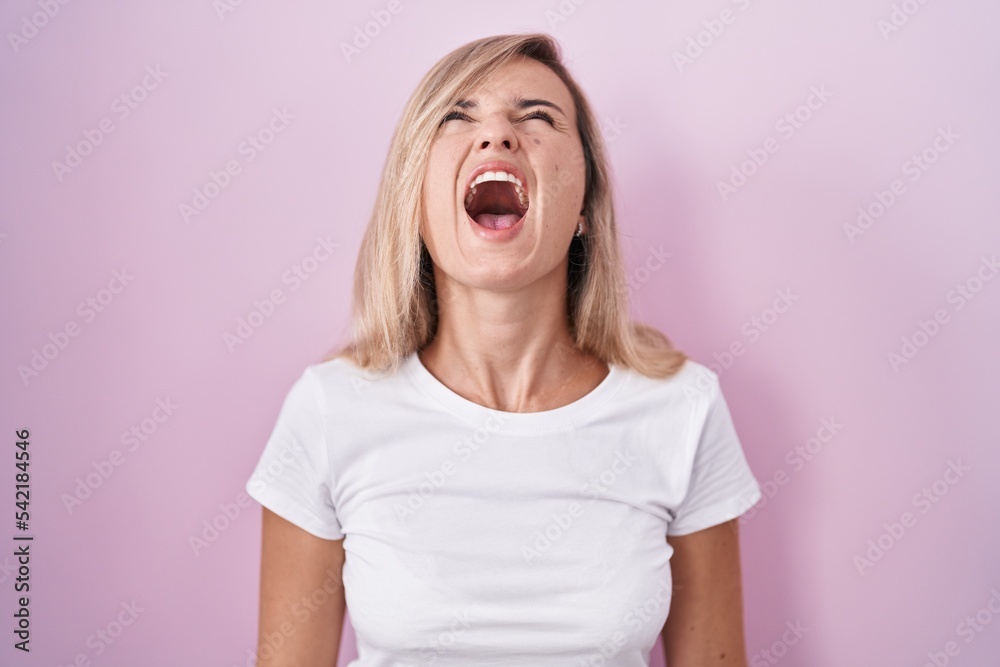 Young blonde woman standing over pink background angry and mad screaming frustrated and furious, shouting with anger. rage and aggressive concept. Young blonde woman standing over pink background angry and mad screaming frustrated and furious, shouting with anger. rage and aggressive concept.