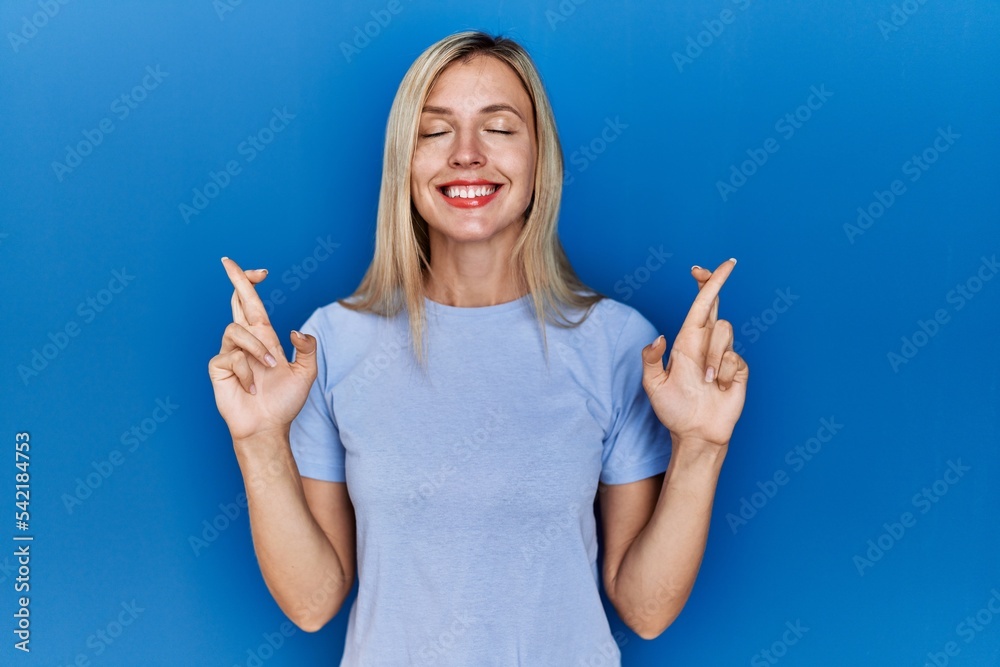 Beautiful blonde woman wearing casual t shirt over blue background gesturing finger crossed smiling with hope and eyes closed. luck and superstitious concept. Beautiful blonde woman wearing casual t shirt over blue background gesturing finger crossed smiling with hope and eyes closed. luck and superstitious concept.
