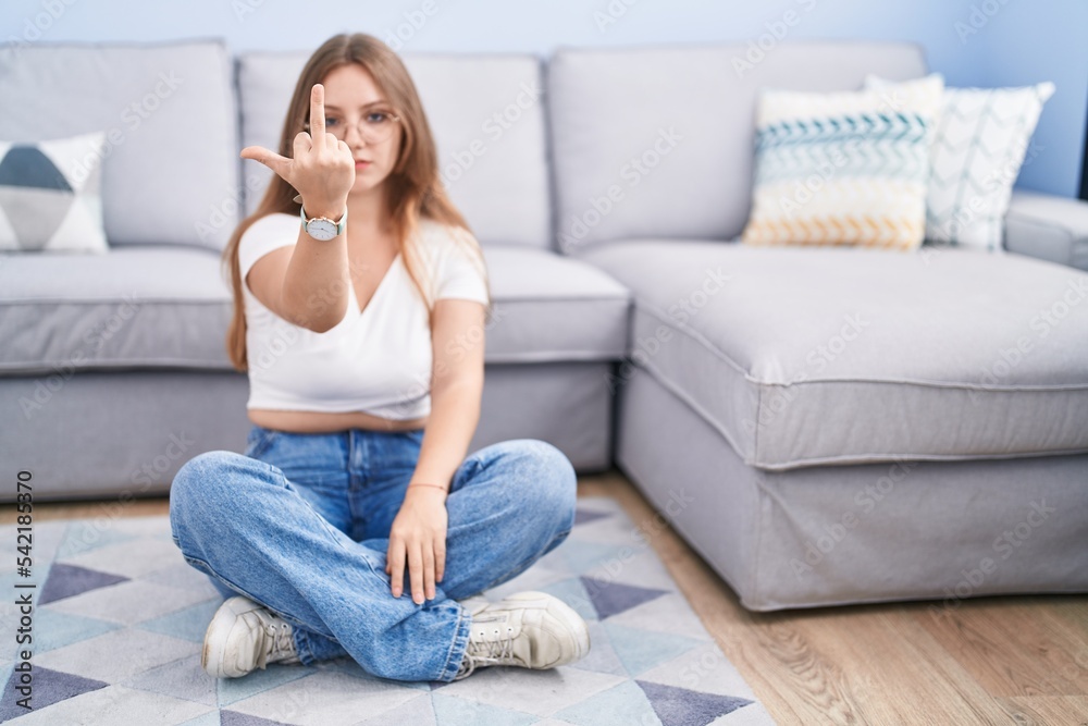 Young caucasian woman sitting on the floor at the living room showing middle finger, impolite and rude fuck off expression Young caucasian woman sitting on the floor at the living room showing middle finger, impolite and rude fuck off expression