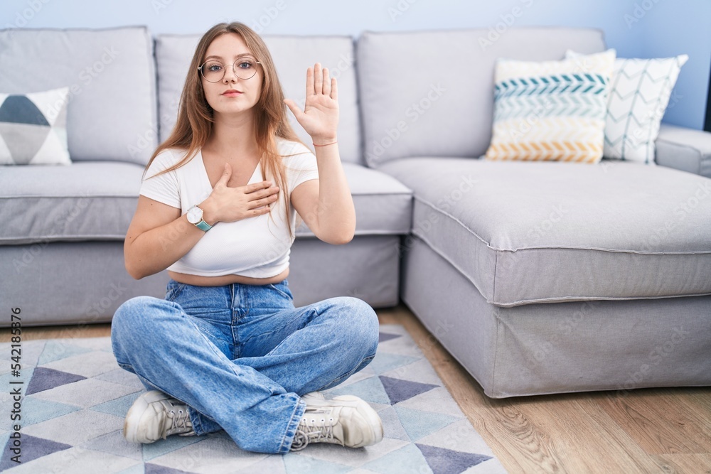 Young caucasian woman sitting on the floor at the living room swearing with hand on chest and open palm, making a loyalty promise oath Young caucasian woman sitting on the floor at the living room swearing with hand on chest and open palm, making a loyalty promise oath