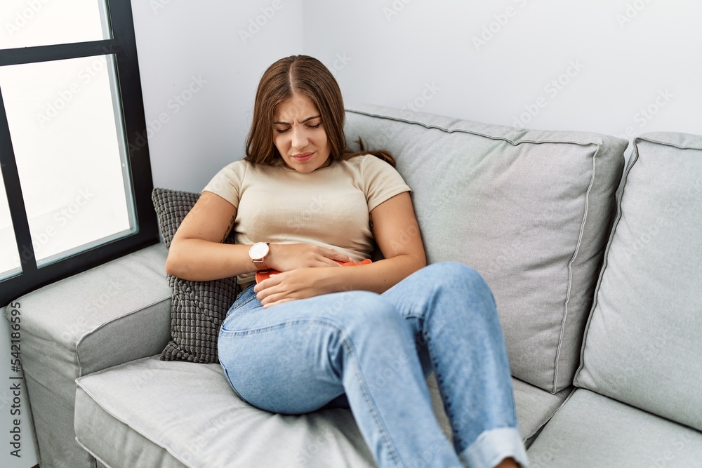 Young hispanic woman using hot water bag for stomachache at home