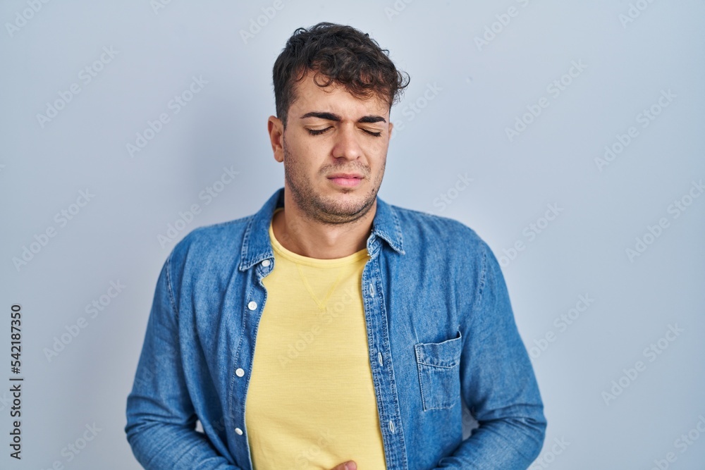 Young hispanic man standing over blue background with hand on stomach because indigestion, painful illness feeling unwell. ache concept. Young hispanic man standing over blue background with hand on stomach because indigestion, painful illness feeling unwell. ache concept.