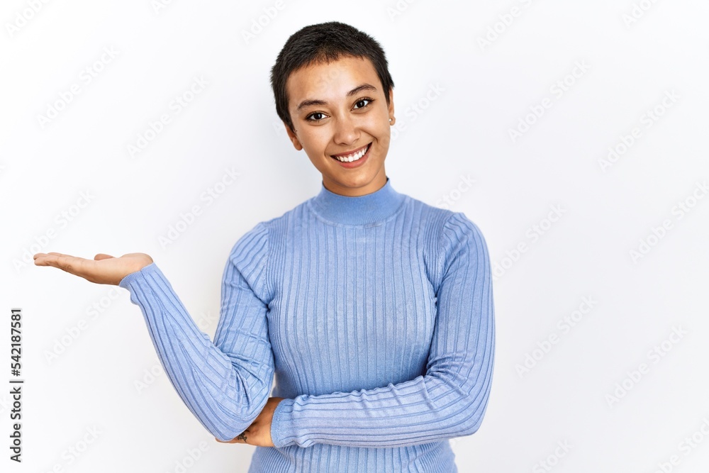 Young hispanic woman with short hair standing over isolated background smiling cheerful presenting and pointing with palm of hand looking at the camera. Young hispanic woman with short hair standing over isolated background smiling cheerful presenting and pointing with palm of hand looking at the camera.