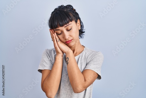 Young hispanic woman wearing casual t shirt over blue background sleeping tired dreaming and posing with hands together while smiling with closed eyes.