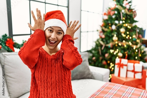 Young hispanic woman with short hair wearing christmas hat sitting on the sofa smiling cheerful playing peek a boo with hands showing face. surprised and exited photo