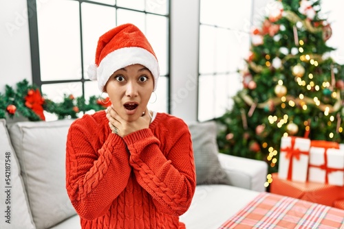 Young hispanic woman with short hair wearing christmas hat sitting on the sofa shouting suffocate because painful strangle. health problem. asphyxiate and suicide concept.