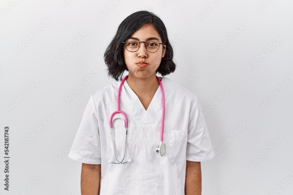 Young hispanic doctor woman wearing stethoscope over isolated background puffing cheeks with funny face. mouth inflated with air, crazy expression. Young hispanic doctor woman wearing stethoscope over isolated background puffing cheeks with funny face. mouth inflated with air, crazy expression.