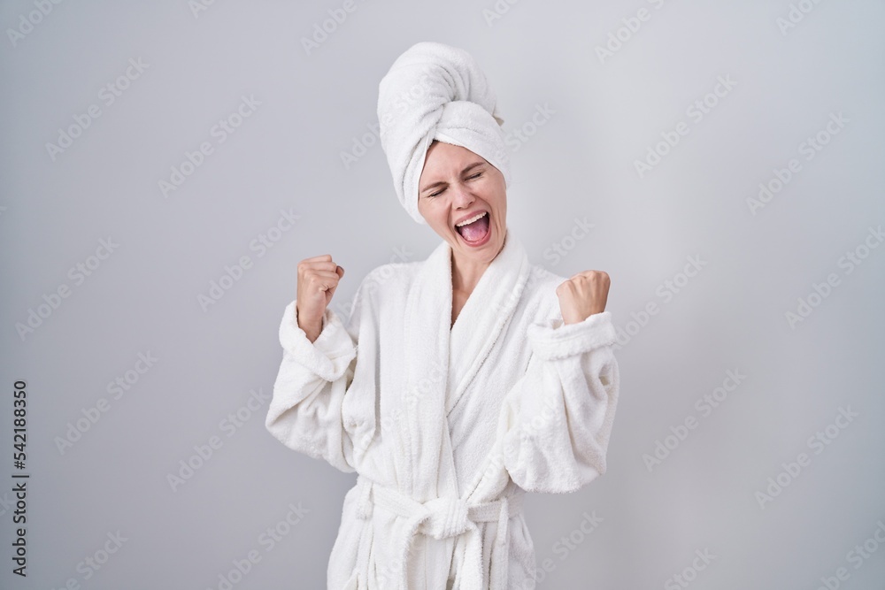 Blonde caucasian woman wearing bathrobe very happy and excited doing winner gesture with arms raised, smiling and screaming for success. celebration concept. Blonde caucasian woman wearing bathrobe very happy and excited doing winner gesture with arms raised, smiling and screaming for success. celebration concept.