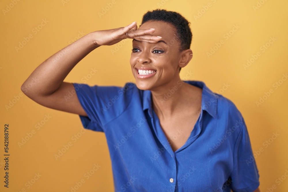 African american woman standing over yellow background very happy and smiling looking far away with hand over head. searching concept.