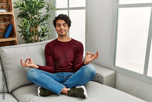 Young hispanic man meditation doing yoga sitting on the sofa at home.