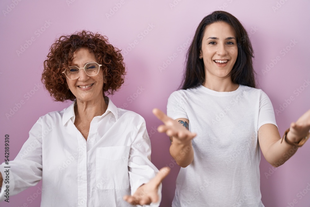 Hispanic mother and daughter wearing casual white t shirt over pink background smiling cheerful offering hands giving assistance and acceptance. Hispanic mother and daughter wearing casual white t shirt over pink background smiling cheerful offering hands giving assistance and acceptance.