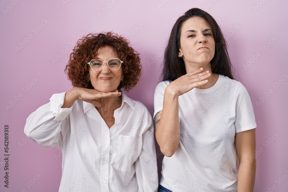 Hispanic mother and daughter wearing casual white t shirt over pink background cutting throat with hand as knife, threaten aggression with furious violence Hispanic mother and daughter wearing casual white t shirt over pink background cutting throat with hand as knife, threaten aggression with furious violence