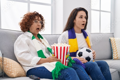 Hispanic mother and daughter watching football supporting team scared and amazed with open mouth for surprise, disbelief face
