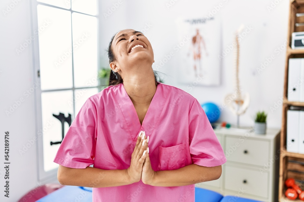 Young hispanic woman working at pain recovery clinic begging and praying with hands together with hope expression on face very emotional and worried. begging. Young hispanic woman working at pain recovery clinic begging and praying with hands together with hope expression on face very emotional and worried. begging.