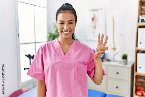 Young hispanic woman working at pain recovery clinic showing and pointing up with fingers number three while smiling confident and happy.